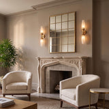 A transitional living room featuring Amarael wall sconces in gold leaf and cloud glass flanking a grid mirror above a carved stone fireplace mantle and textured ivory armchairs.