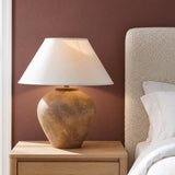 A transitional bedroom with the Welles Table Lamp 23", Distressed Brown with White Shade set on an oak nightstand beside a textured white bouclé headboard.