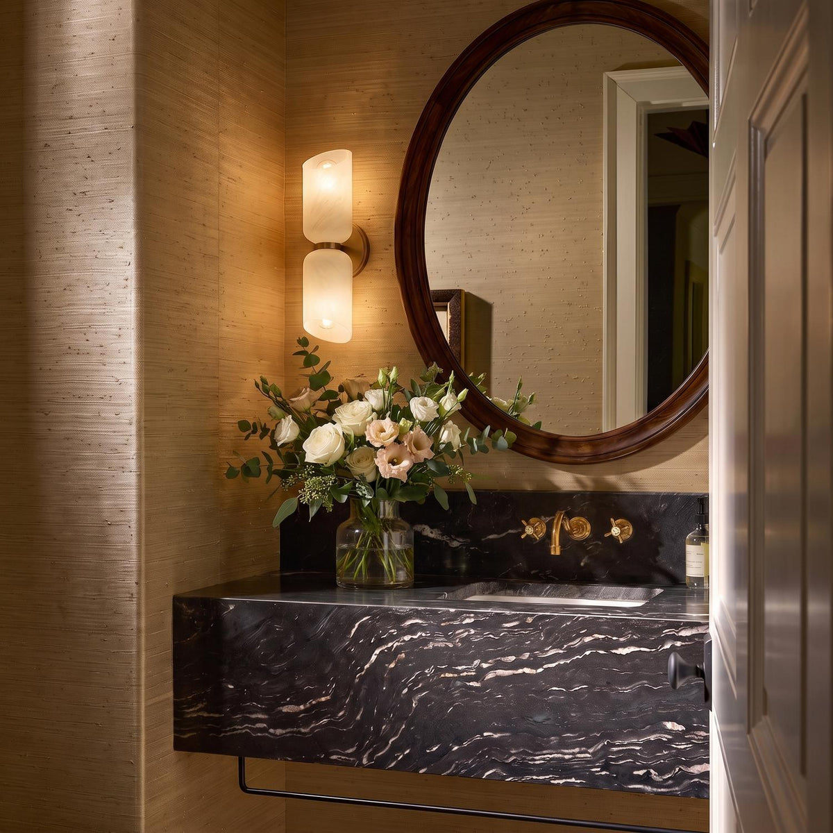 A transitional bathroom with a patina brass and cloud glass wall sconce mounted on grasscloth wallpaper beside a round wood-framed mirror and black marble vanity.