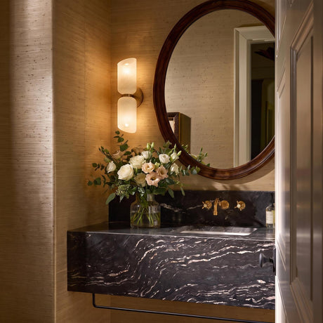 A transitional bathroom with a patina brass and cloud glass wall sconce mounted on grasscloth wallpaper beside a round wood-framed mirror and black marble vanity.