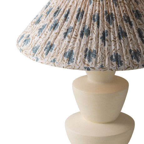 A close-up of a Bonnie Table Lamp with a pleated fabric shade featuring a blue floral and gold vine pattern on a cream background, paired with a matte cream-colored ceramic base.