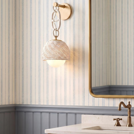 A coastal bathroom with the Kairo Wall Sconce in champagne leaf and bleached rattan mounted on blue striped wallpaper beside a brass-framed mirror over a marble vanity.