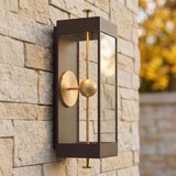 A modern home exterior featuring a Granville LED Outdoor Wall Light in textured bronze and patina brass mounted on a tan stone wall with golden autumn foliage.
