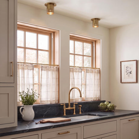 A transitional kitchen with two patina brass Bevan Mini Cylinder flush mounts over a dark marble counter with wood-framed windows and white cafe curtains.