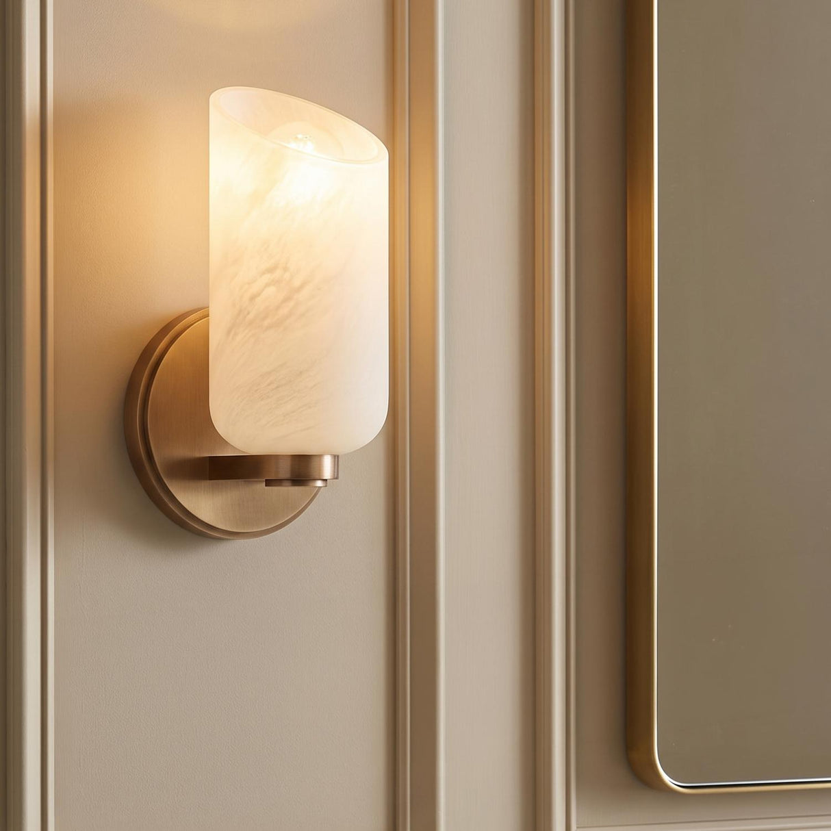 A transitional bathroom featuring the Vivara Wall Sconce with patina brass and cloud glass mounted on a paneled beige wall beside a matching brass-framed mirror.