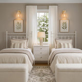 A transitional bedroom featuring two Birdie Wall Sconce 13", Gold Leaf and Natural Rope fixtures mounted above twin white iron beds flanking a central window and nightstand.