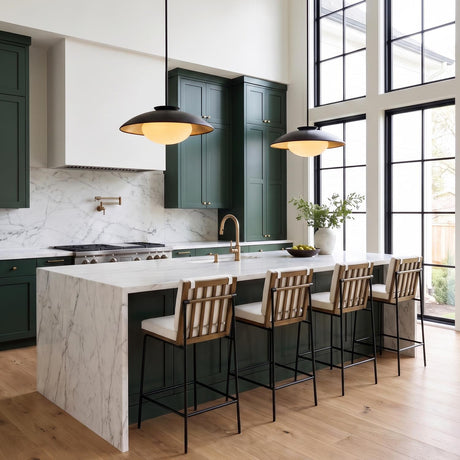 A contemporary kitchen with two matte black and gold leaf saucer pendants over a white marble waterfall island, featuring forest green cabinets and double-height windows.