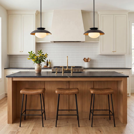 A modern kitchen with two matte black and gold leaf Vera Medium Pendants suspended over a black stone island with natural wood and leather barstools.