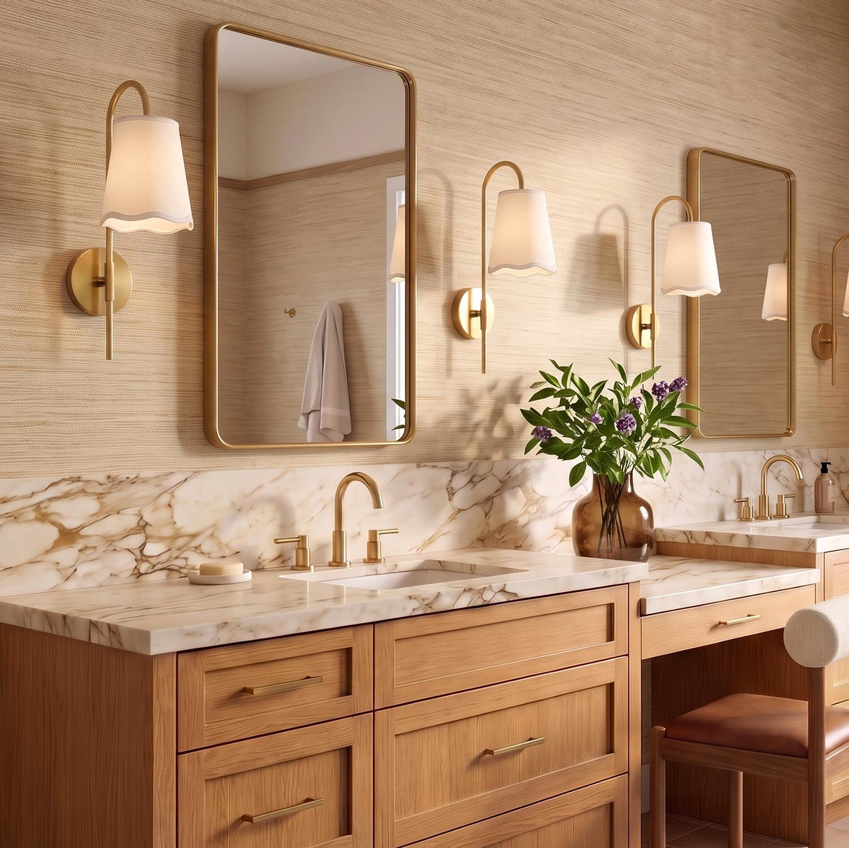 A transitional bathroom with aged brass wall sconces featuring white linen shades flanking rectangular brass mirrors above a marble vanity and wood cabinets with textured grasscloth wallpaper.