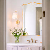 A transitional bathroom featuring a Basil Wall Sconce 19", Aged Brass with White Shade mounted beside a gold scalloped mirror above a white marble vanity with brass faucet.