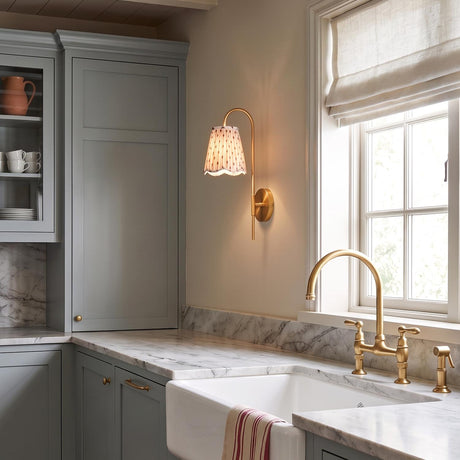 A farmhouse kitchen featuring an aged brass wall sconce with a printed shade beside a window above marble counters and a white sink under a beamed ceiling.