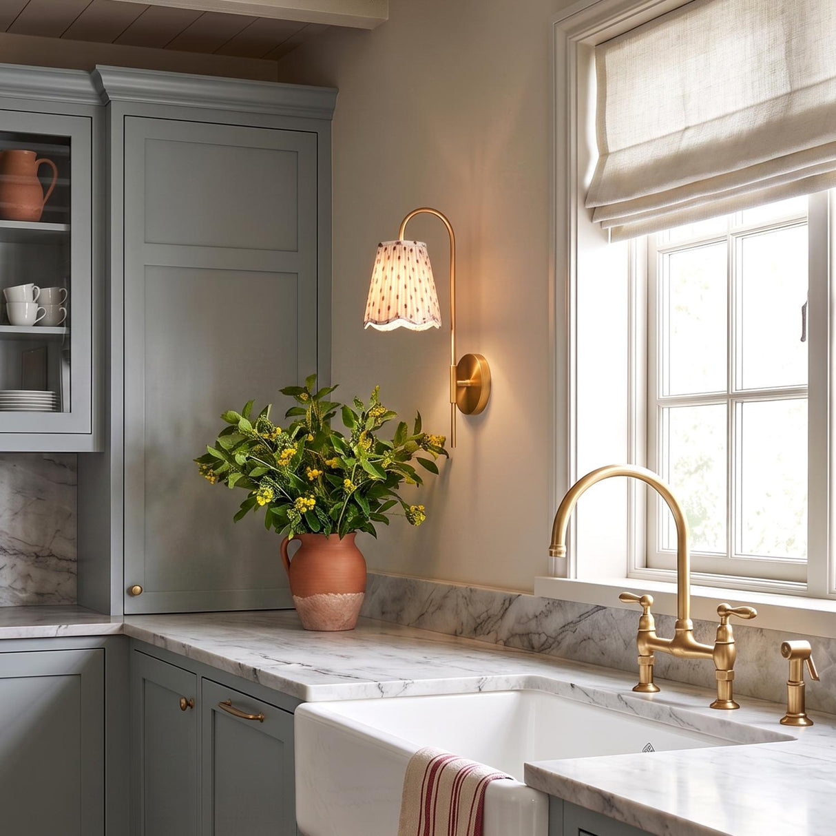 A modern farmhouse kitchen featuring the Basil Wall Sconce in aged brass with a red primrose printed shade mounted over marble countertops beside a window.
