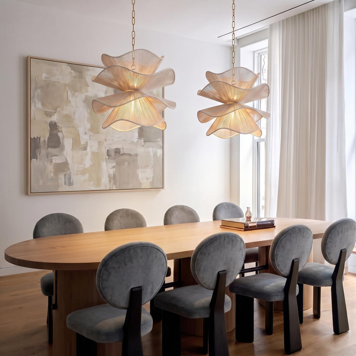 A modern dining room featuring two Lotus Pendants in aged brass and natural cotton suspended over a light wood oval table beside a large abstract painting.