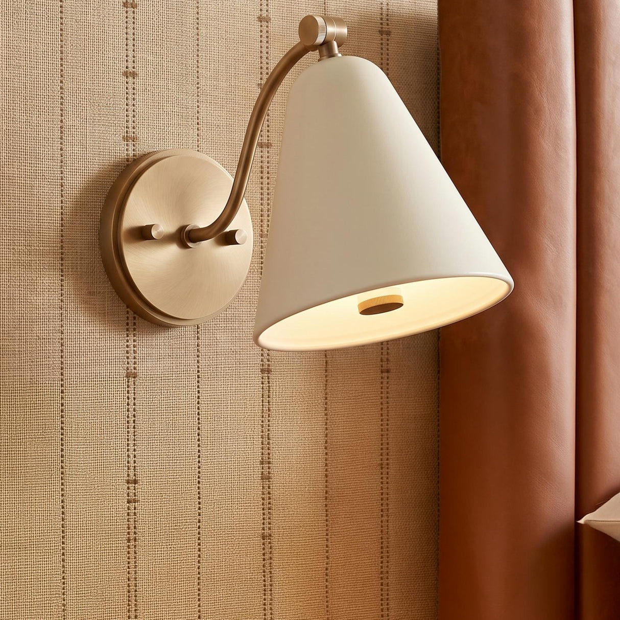 A transitional bedroom with a patina brass and soft sand Bryden Pivot Wall Sconce mounted on striped beige wallpaper beside a brown leather headboard.