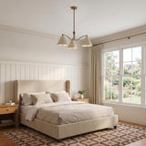 A transitional bedroom featuring the Bryden 3 Light Chandelier in patina brass and soft sand centered above a beige upholstered bed and white vertical wainscoting.