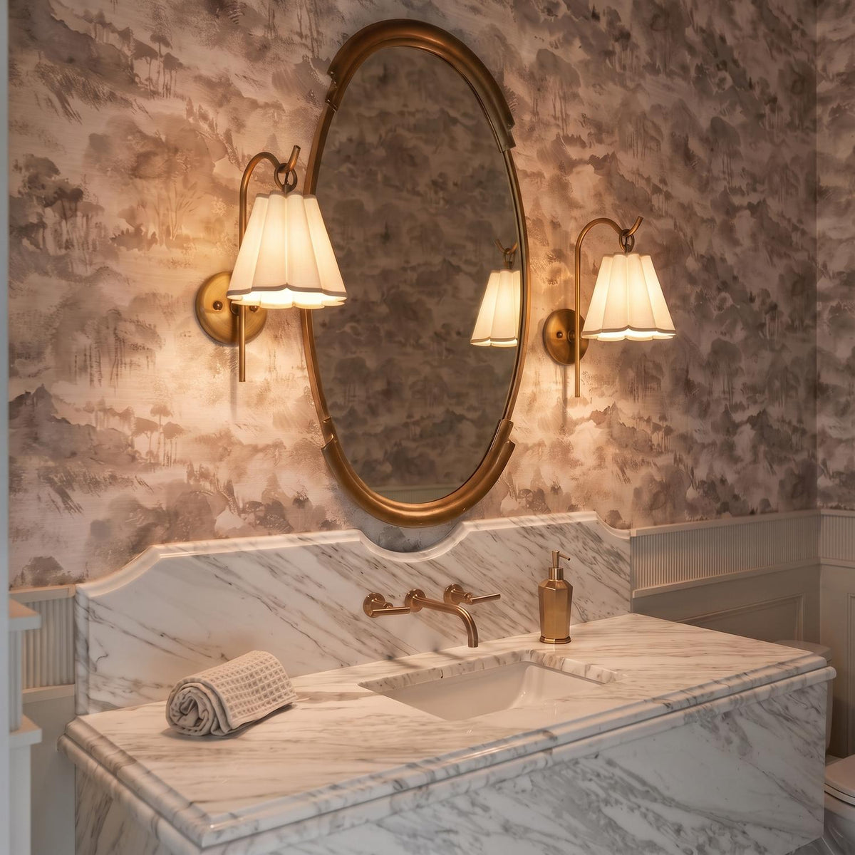 A traditional bathroom with two patina brass wall sconces featuring pleated linen shades flanking an oval mirror above a white marble vanity with scenic wallpaper.