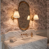 A traditional bathroom with two patina brass wall sconces featuring pleated linen shades flanking an oval mirror above a white marble vanity with scenic wallpaper.