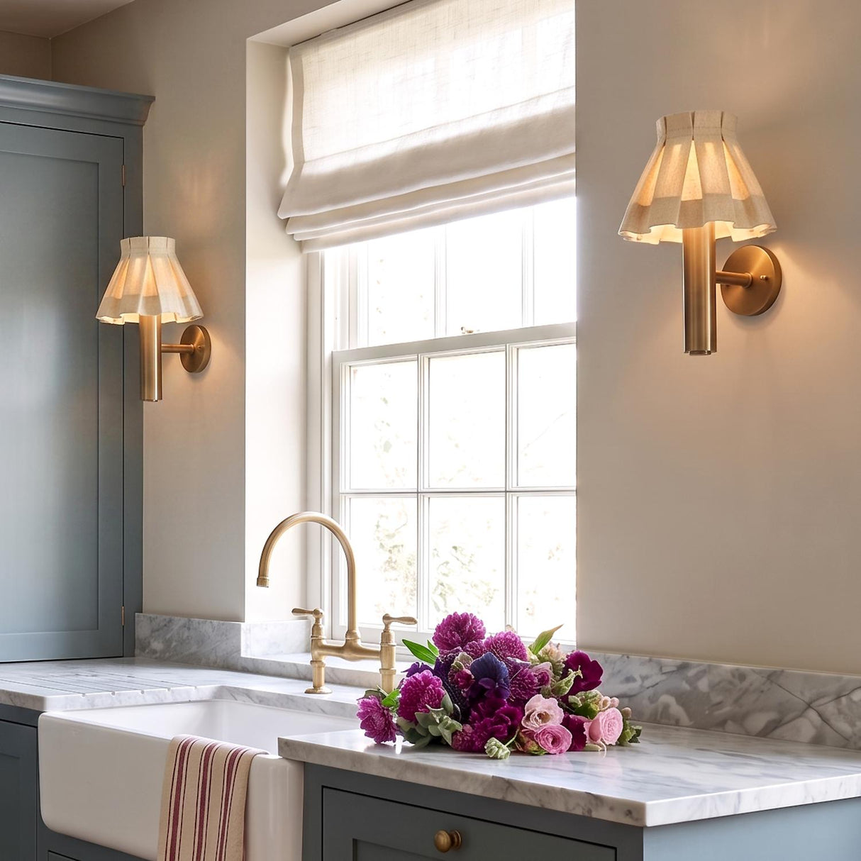 A transitional kitchen with two Tabitha wall sconces in patina brass with wheat pleated linen shades flanking a window over a marble counter and apron-front sink.
