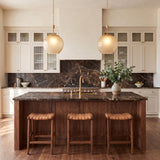 A transitional kitchen with two Annara Medium Pendants in aged brass and champagne frosted glass hanging over a walnut island with dark marble and woven barstools.
