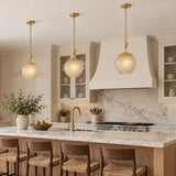 A transitional kitchen features three Annara Small Pendants in aged brass and champagne frosted glass hanging above a marble island surrounded by woven wooden barstools.
