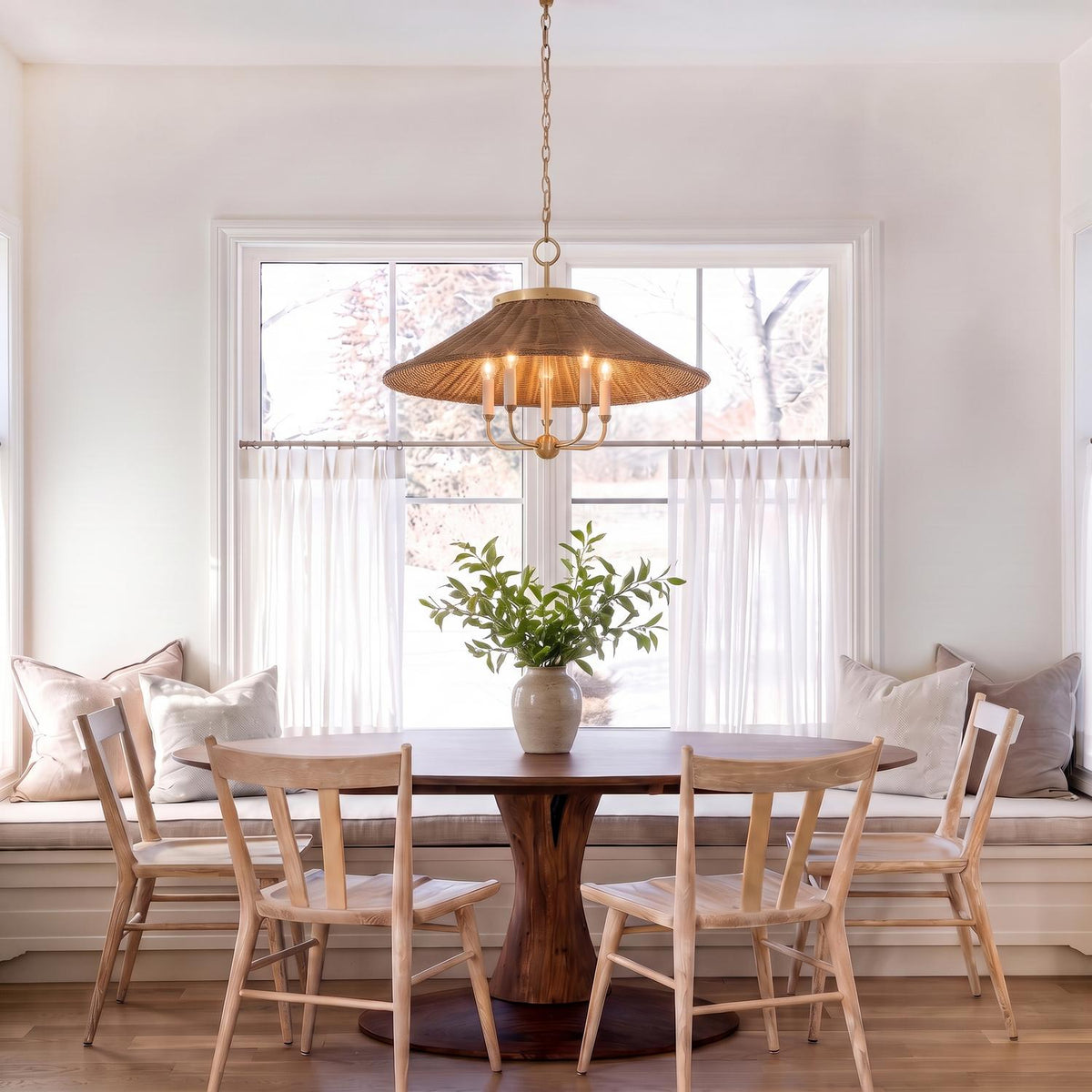 A transitional dining room with a woven wicker and aged brass chandelier hanging over a round wood table beside a window bench.