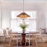 A transitional dining room with a woven wicker and aged brass chandelier hanging over a round wood table beside a window bench.