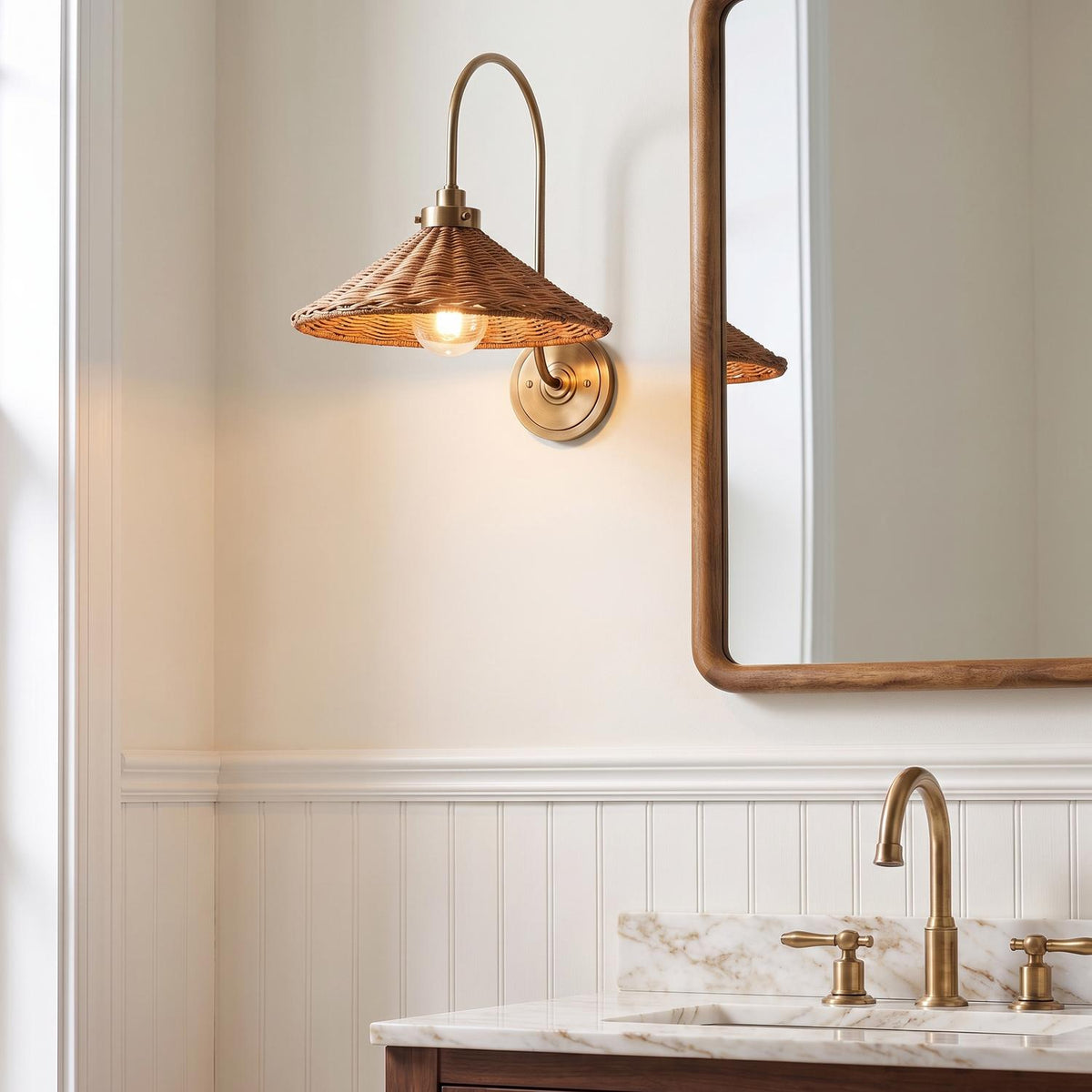 A coastal bathroom featuring the Bramble Wall Sconce in walnut wicker and aged brass mounted beside a wood-framed mirror above a marble vanity with beadboard.