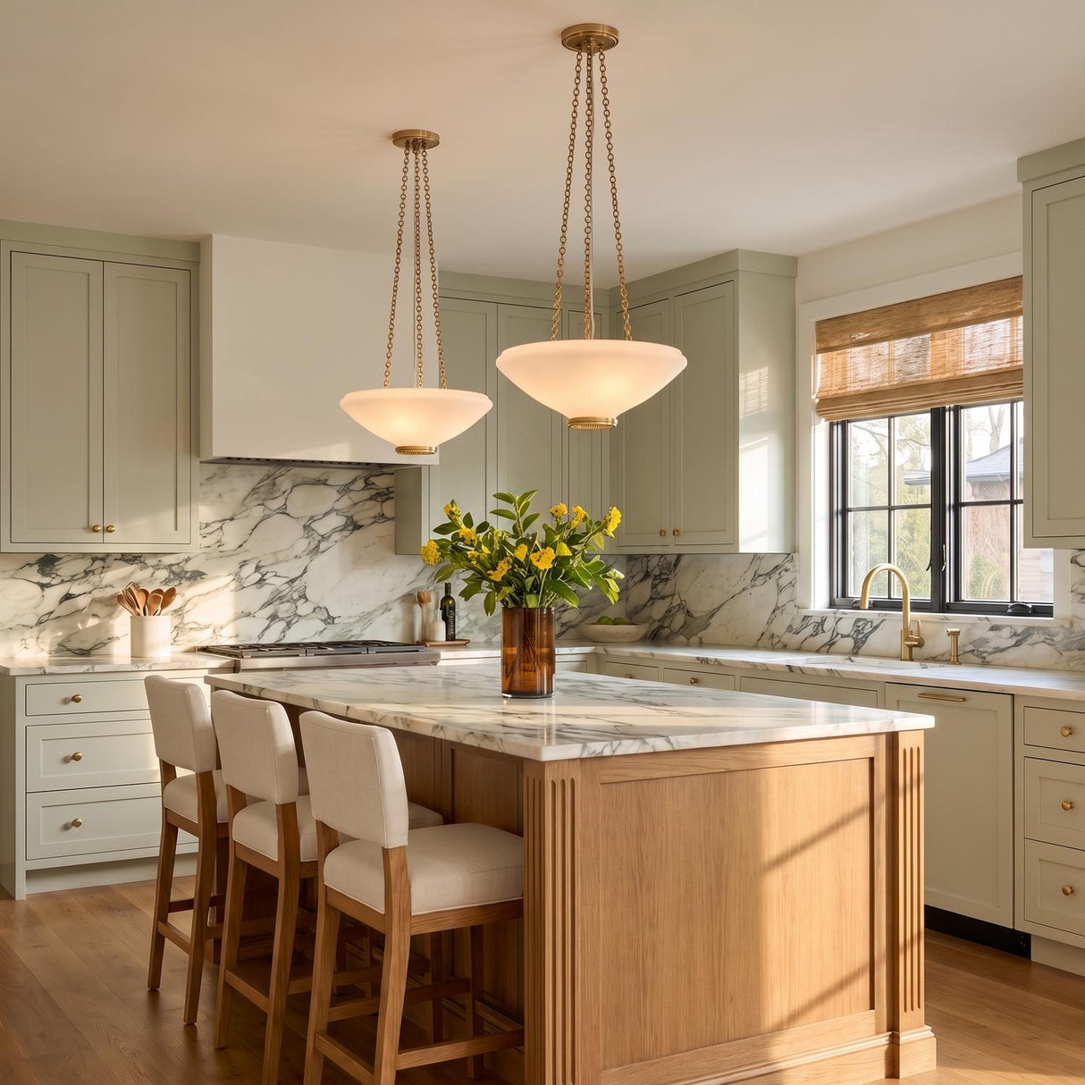 A transitional kitchen with two Lindor Medium Pendants in aged brass and opal glass hanging over a white marble island and sage green cabinetry.