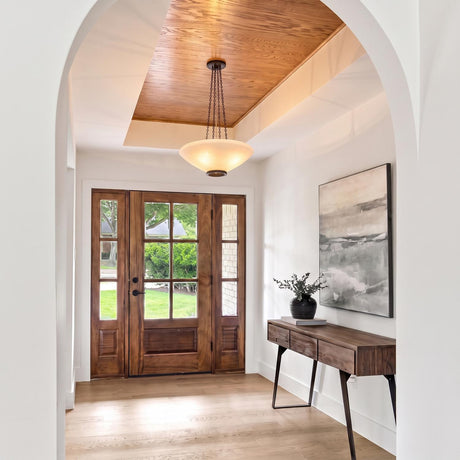 A transitional foyer with a distressed bronze and opal glass pendant hanging from a wood-paneled tray ceiling above a walnut console table and wood front door.
