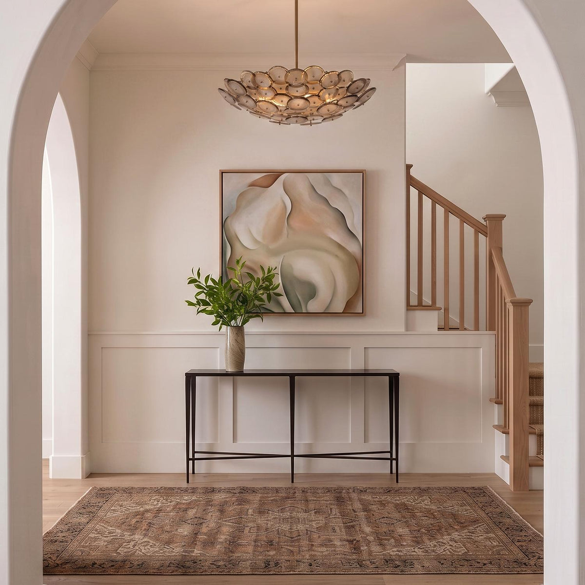 A transitional foyer with a patina brass and smoke taupe piastra glass chandelier centered above a dark console table, white wainscoting, and a light wood staircase.