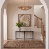 A transitional foyer with a patina brass and smoke taupe piastra glass chandelier centered above a dark console table, white wainscoting, and a light wood staircase.