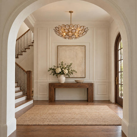 A transitional foyer featuring the Perenna Oversized Chandelier with patina brass accents and smoke taupe piastra glass suspended above a dark wood console table and staircase.