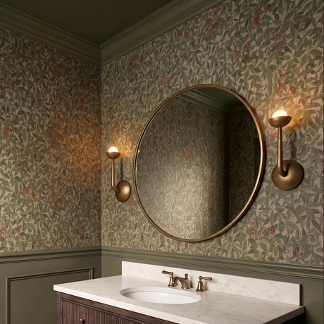 A traditional bathroom featuring two Bettina LED Wall Sconce 12 inch Vintage Brass fixtures flanking a round mirror over a white marble vanity and green floral wallpaper.