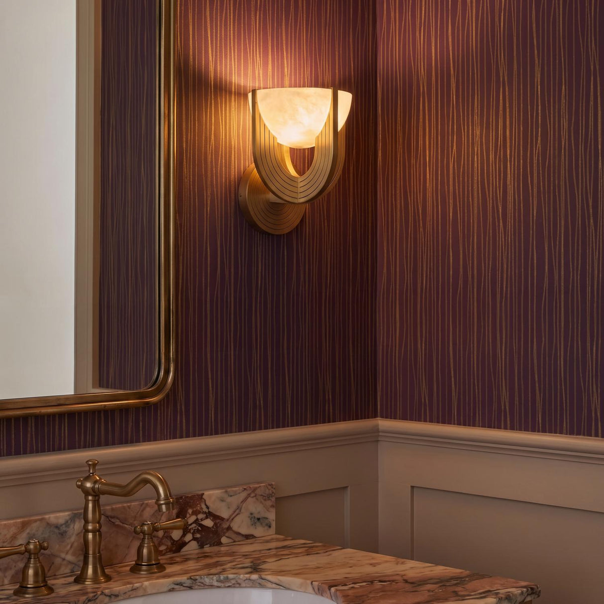 A transitional bathroom featuring an Armand LED Wall Sconce 8", Vintage Brass and Alabaster, mounted to plum-colored textured wallpaper above a pink marble countertop and brass faucet.
