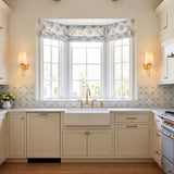 A traditional kitchen with two gold leaf wall sconces featuring raw silk shades flanking a large bay window above a white farmhouse sink and patterned tile.
