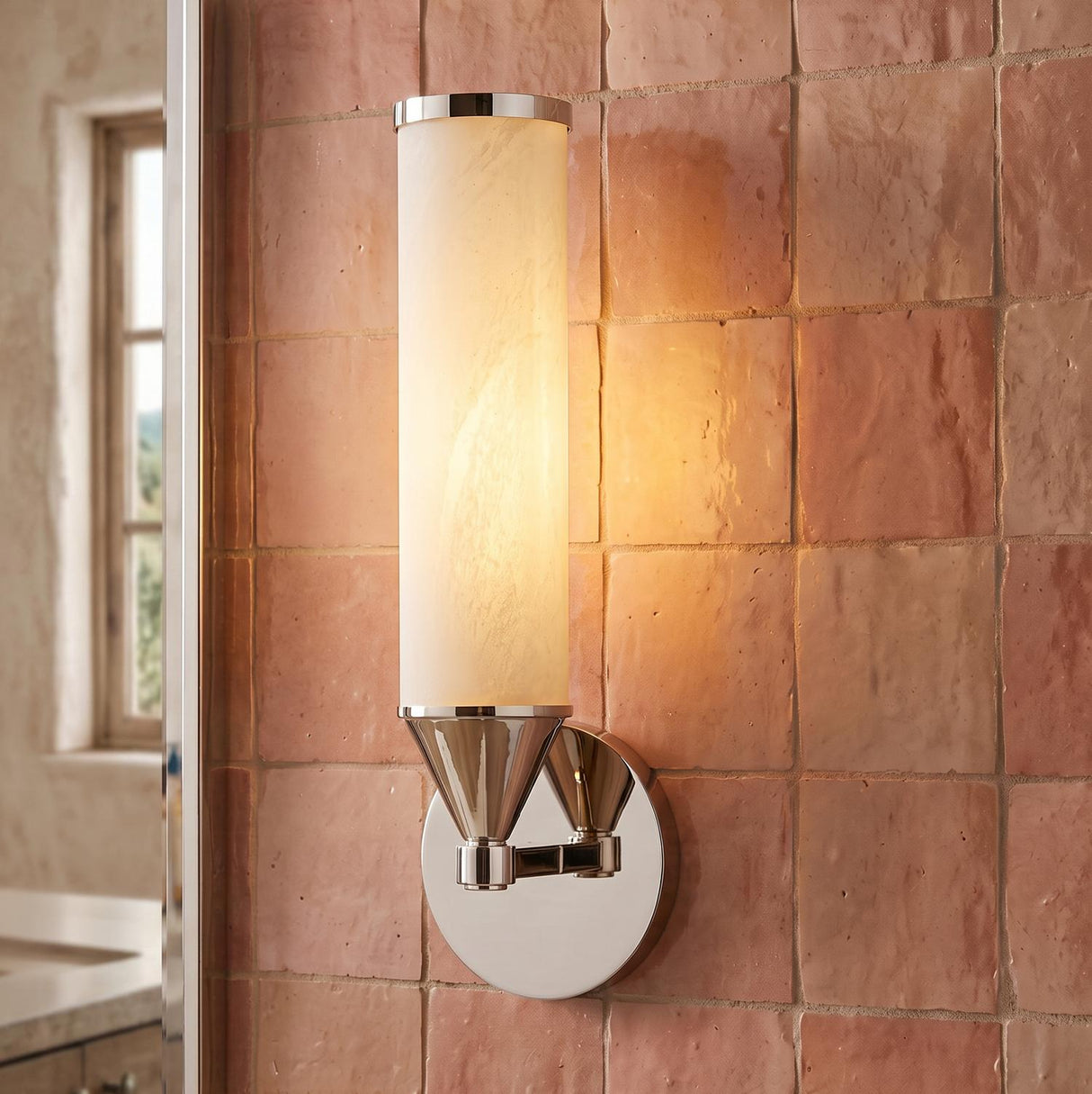 A Mediterranean bathroom with a Sayer Wall Sconce 15", Polished Nickel and Cloud Glass mounted to pink Zellige tiles beside a mirror above a marble vanity.