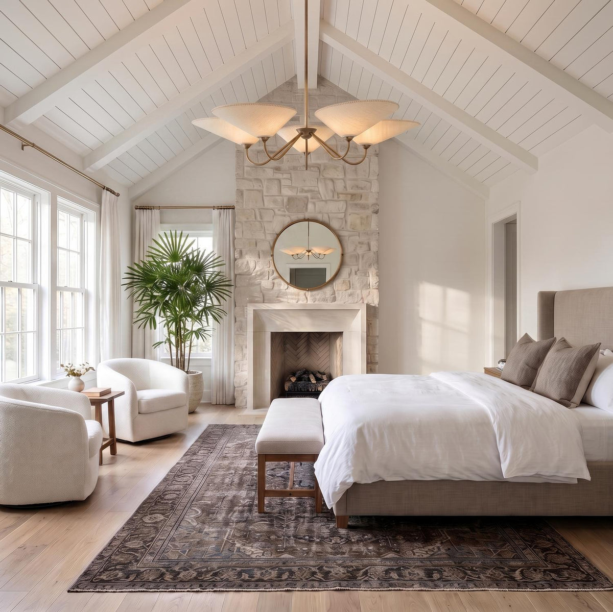 A modern farmhouse bedroom with the Zayla 5 Light Chandelier in patina brass and textured linen centered on a vaulted beamed ceiling above a stone fireplace.