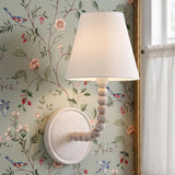 A traditional bedroom featuring the Casimir Curved Wall Sconce 13", Matte Chalk with white wood beads and a linen shade mounted on sage green floral bird wallpaper.