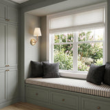 A transitional bedroom with a gilded brass wall sconce and linen shade mounted beside a sage green window seat featuring striped cushions and charcoal pillows.