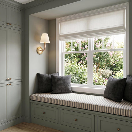 A transitional bedroom with a gilded brass wall sconce and linen shade mounted beside a sage green window seat featuring striped cushions and charcoal pillows.