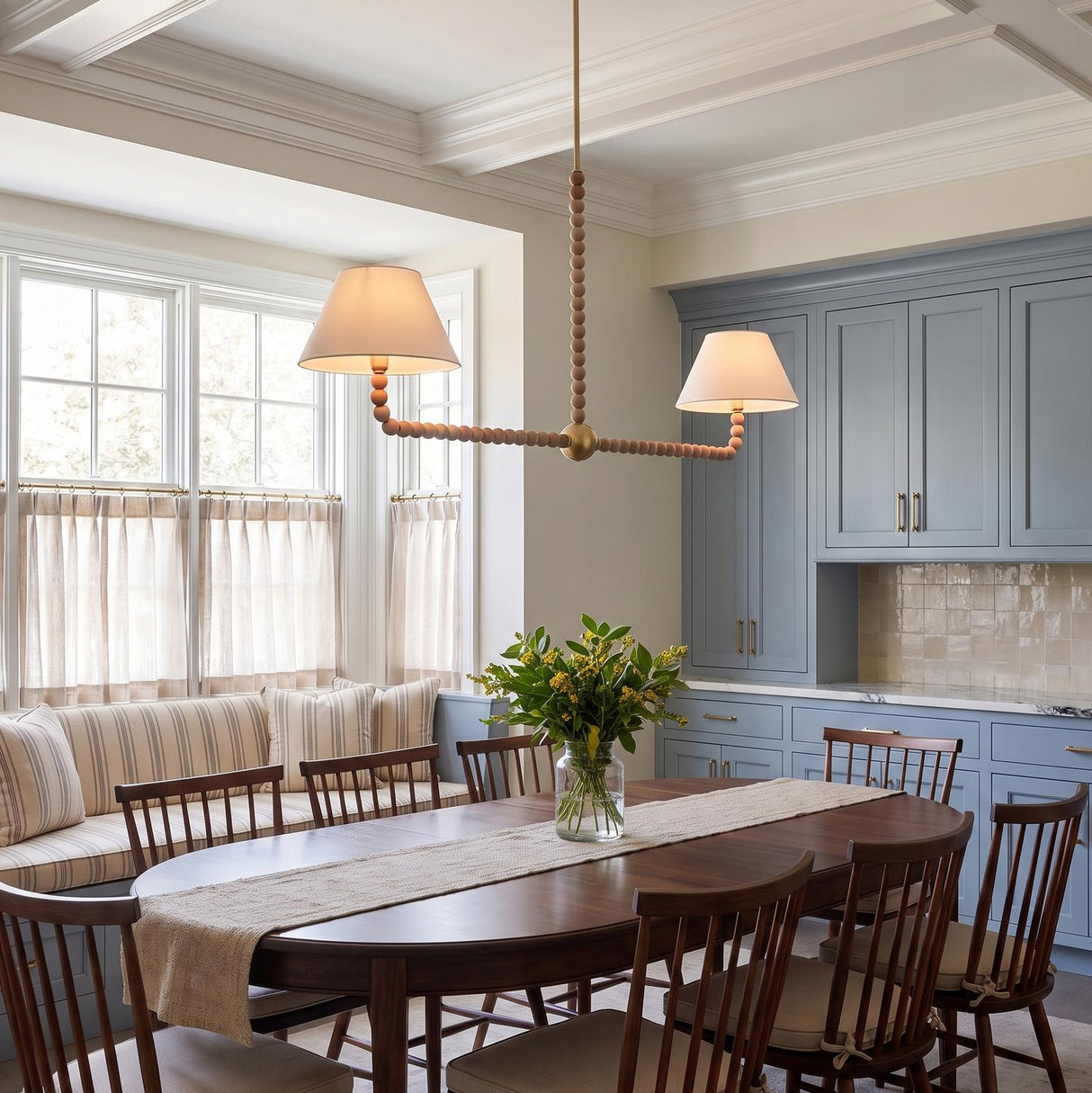 A transitional dining room featuring a linear pendant with beaded wood and linen shades over a dark wood table beside blue cabinetry and coffered ceilings.