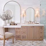 A transitional bathroom with two Casimir Wall Sconces in Matte Chalk flanking a brass arched mirror above a fluted wood vanity and marble hexagon tile floor.