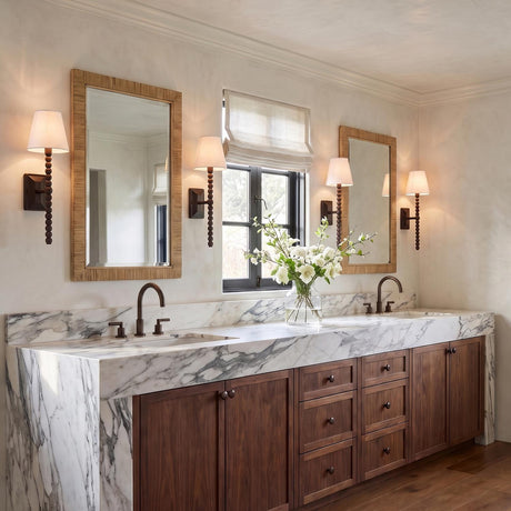 A transitional bathroom with aged iron wall sconces featuring white linen shades flanking two mirrors over a marble vanity with walnut cabinetry and bronze faucets.