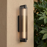 A modern home exterior featuring a Halden Matte Black and Smoke Glass LED Wall Sconce glowing on a beige stucco pillar beside green leafy plants at dusk.