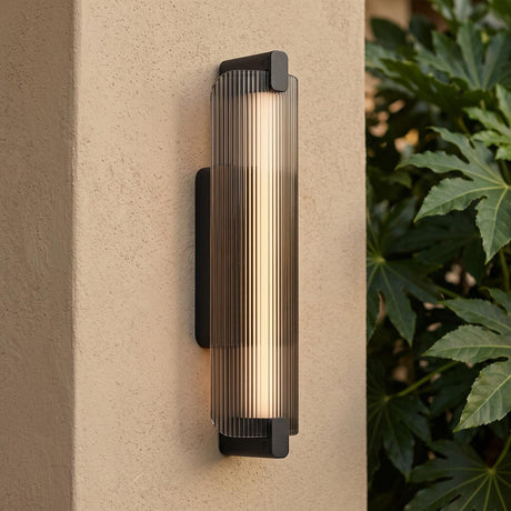 A modern home exterior featuring a Halden Matte Black and Smoke Glass LED Wall Sconce glowing on a beige stucco pillar beside green leafy plants at dusk.
