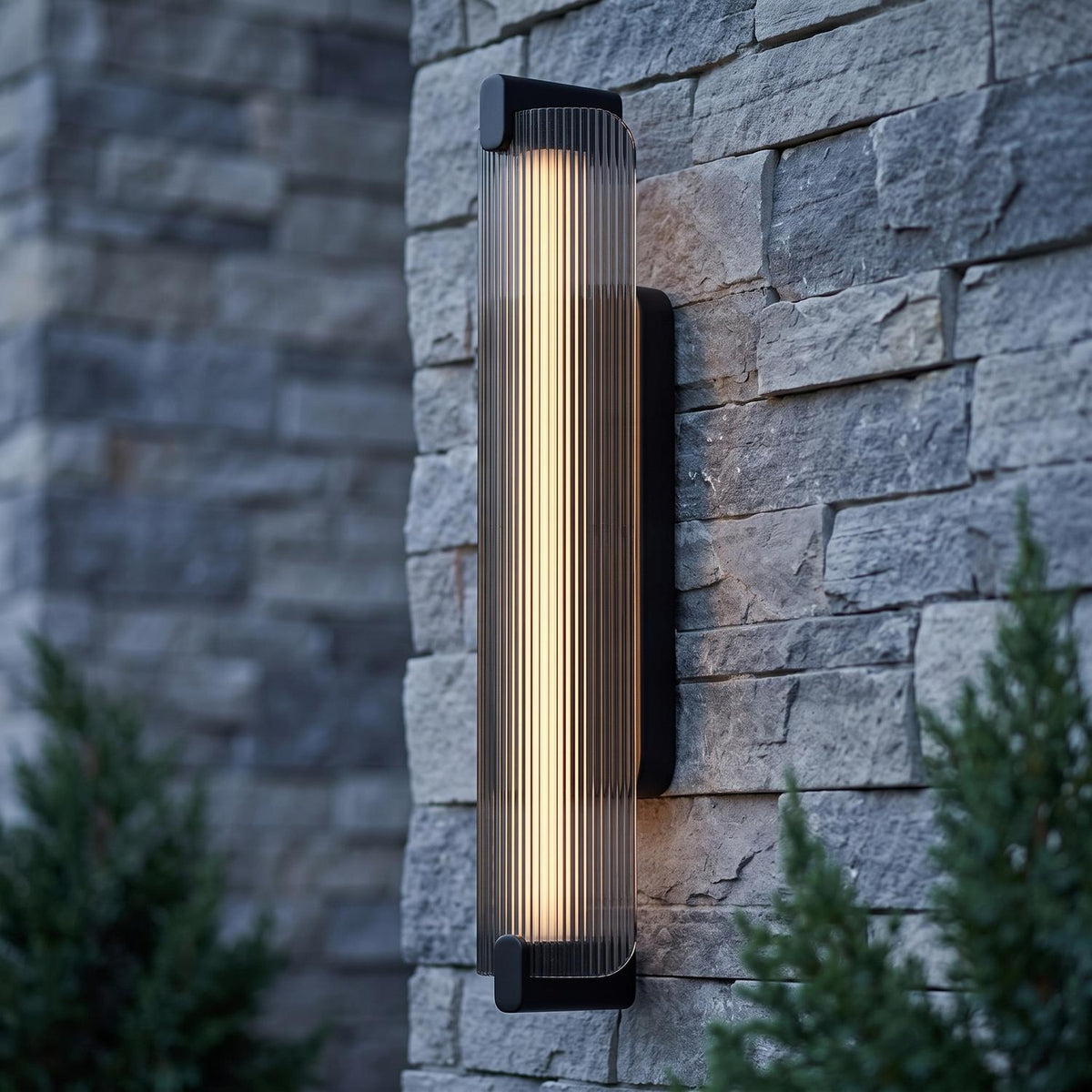 A modern home exterior featuring the Halden LED wall sconce in matte black and smoke glass mounted on a gray stone pillar at twilight near evergreen bushes.