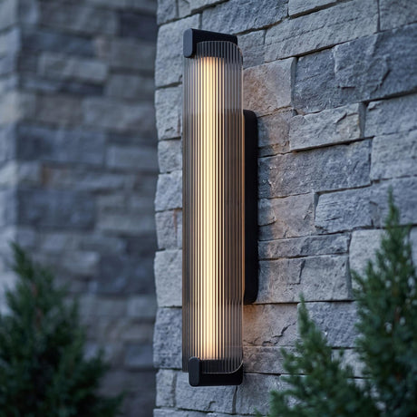 A modern home exterior featuring the Halden LED wall sconce in matte black and smoke glass mounted on a gray stone pillar at twilight near evergreen bushes.