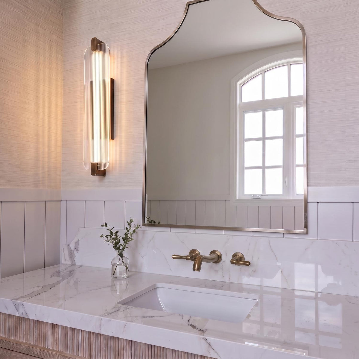 A transitional bathroom with the Halden LED Wall Sconce in weathered brass and ribbed glass mounted beside a curved mirror above a white marble countertop.