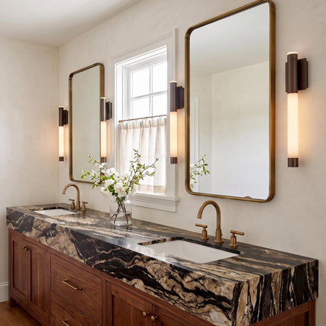 A transitional bathroom features Anders Large LED Wall Sconce 24-inch Pewter Bronze and Alabaster light fixtures mounted beside brass mirrors over a veined marble countertop.