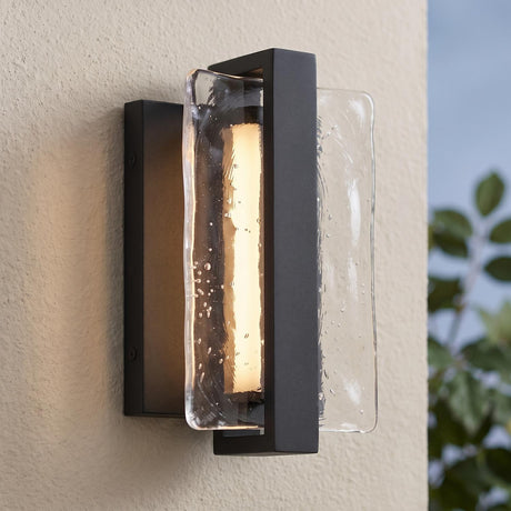 A modern home exterior with textured black Kendrick LED Outdoor Wall Light with seeded cast glass glowing on a beige stucco wall at dusk beside green foliage.